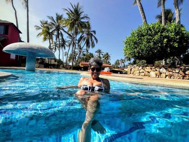 JuliaNewell with small black tits, wearing sunglasses and a black and white bikini, standing in a clear blue pool, surrounded by palm trees and a stone wall.