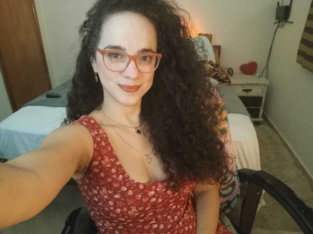 AmyFlores with curly black hair, red glasses, and red floral dress, smiling at the camera, showing her small tits and tattooed arm, sitting in a chair.