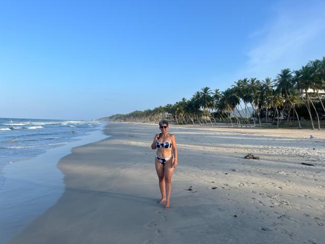 JuliaNewell stands on a beach, wearing a blue and black bikini, her tits covered, ass exposed, brown hair in a bun, sunglasses on, palm trees in the background.