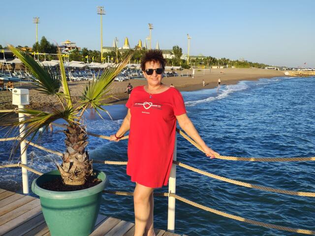 DoraBlueEyess, middle-aged woman with short curly brown hair, wearing red dress and sunglasses, standing by blue water, holding rope, palm tree in green pot, sunny beach.