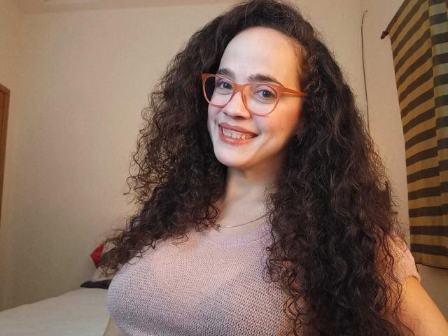 AmyFlores with long curly brown hair, wearing red glasses and a pink knit top, smiling at the camera, showing her tits. She has fair skin and a cute face.