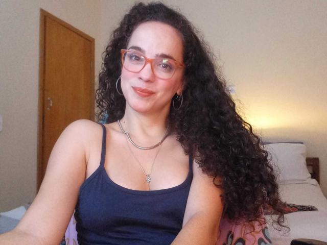 AmyFlores with curly black hair, red glasses, and a black tank top, smiling at the camera, showing her small tits and a silver necklace. Bedroom background.