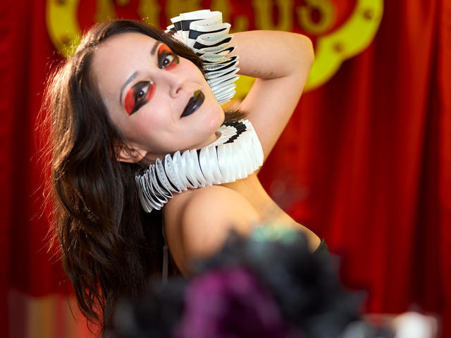 MollyMarmalade with dark makeup, red eyeshadow, black lipstick, wearing a white ruffled choker and zebra-striped gloves, posing with arms up, showing her tits, against red curtain.