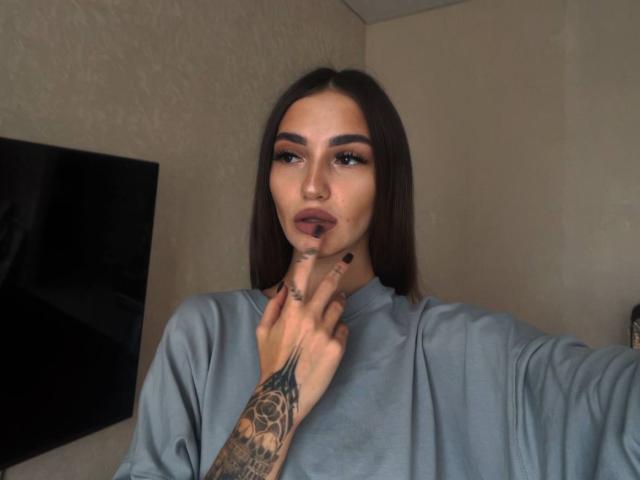 SophieKitty has long brown hair, dark eyebrows, and is wearing a gray shirt, touching her chin with a tattooed finger. She has full lips and dark makeup.