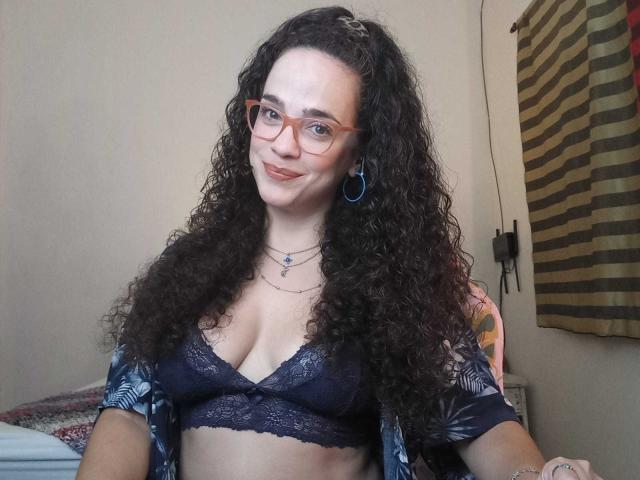 AmyFlores with curly dark hair, wearing glasses, blue lace bra, open shirt, and necklace, smiling at camera, showing her tits, with blue earrings and a tattoo on her arm.