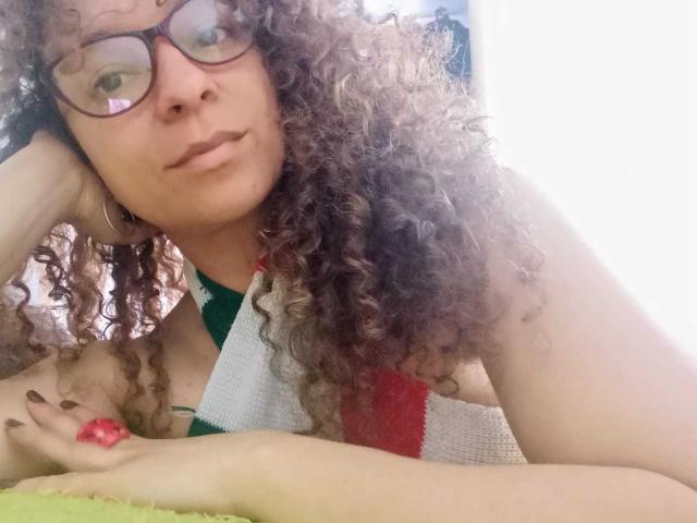 Angelonix with curly brown hair, glasses, and brown lipstick, leaning on arm, wearing green and white top, holding red strawberry, white background. Boobs, ass, pussy not visible.