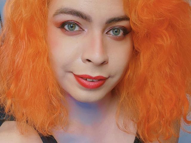 TsFoxCandy has bright orange curly hair, green eyes, red lipstick, and a slight smile, showing a cute, sexy face with subtle makeup and a light skin tone.