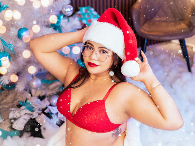 LilyRousee with glasses, red bra, and Santa hat, showing her big boobs, red lipstick, and hand in her hair, snowing in front of a decorated Christmas tree.