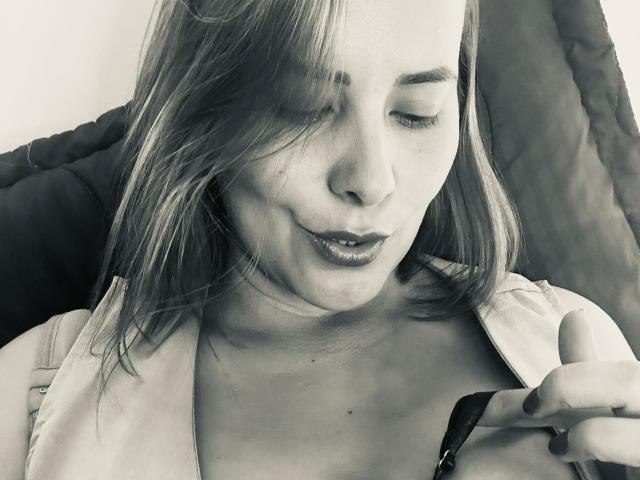 SaritaVelez has long, slightly messy hair, half-closed eyes, and dark lipstick, with a relaxed, slightly smirking expression, sitting against a dark, textured background.
