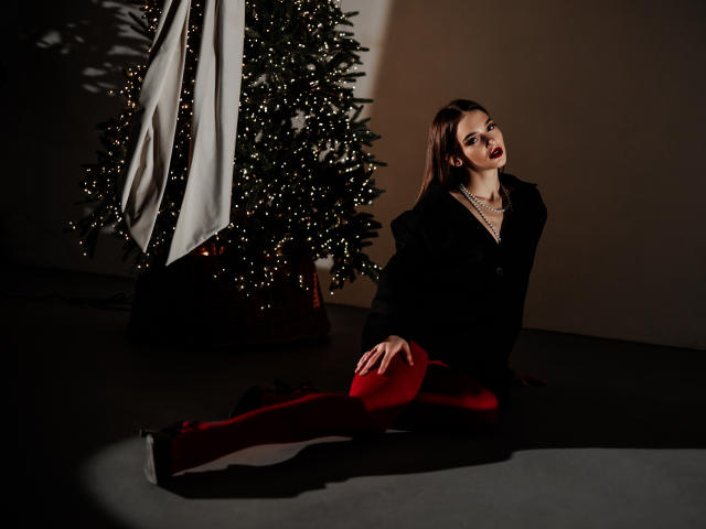 SophieKitty kneeling in front of a Christmas tree with red tights, black jacket, and dark lipstick, looking seductively at the camera, with a shadow on the floor.
