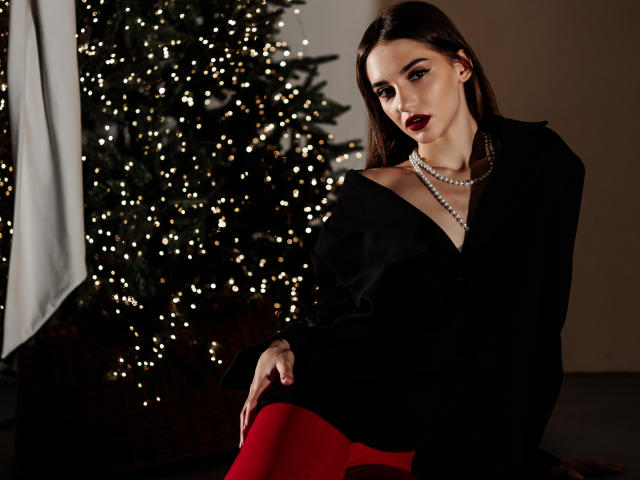 SophieKitty with dark red lipstick, black off-shoulder top, pearl necklace, and red tights, sitting next to a Christmas tree with glowing lights. Her tits are partially visible.