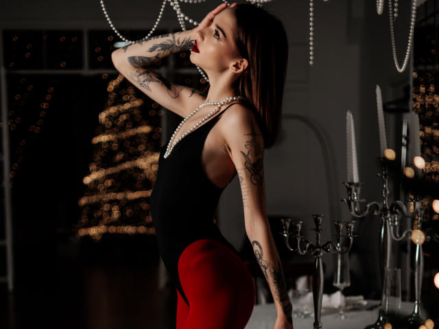 SophieKitty with tattoos, wearing a black top and red pants, posing with hand on head, wearing a pearl necklace, standing by candles, looking up, dark hair, red lipstick.
