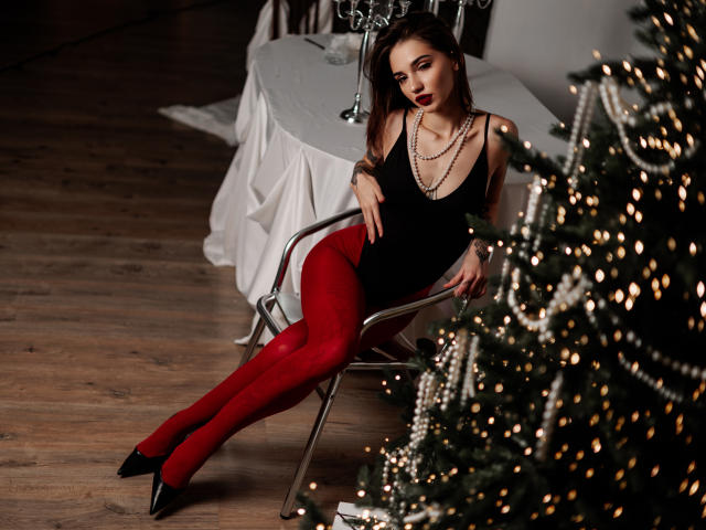 SophieKitty sits on a metal chair wearing a black tank top, red tights, black heels, and a necklace. She's next to a Christmas tree with lights.