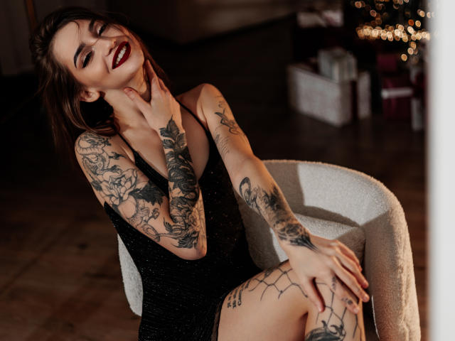 SophieKitty in a black, sparkly dress, sitting in a white chair, smiling with red lipstick, tattoos on arms and legs, brown hair, hand on neck, wood floor.