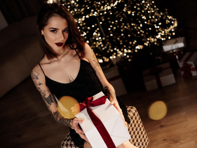 SophieKitty with tattoos, dark lipstick, black sparkly dress, holding gift with red ribbon, yellow balloon, standing in front of Christmas tree with bokeh lights.