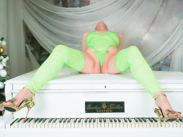 SquirtyCumShow, legs spread, wearing neon green lace lingerie, thigh-high stockings, and gold heels, on a white piano, head tilted back, exposed pussy, visible ass.