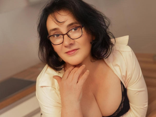 DorisMature with black wavy hair, glasses, and a slightly open white shirt, exposing her large tits and black lace bra, sitting with a soft smile and hand near her neck.