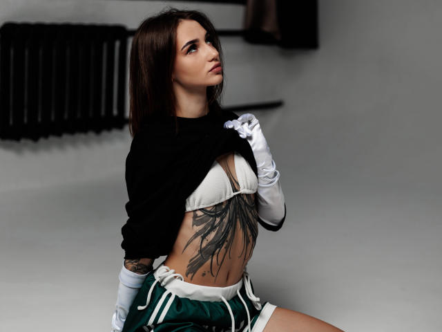 SophieKitty with medium tits, white bra, black sweater, green shorts, white gloves, and a large black tattoo on her flat stomach, looking up with a slight smile.