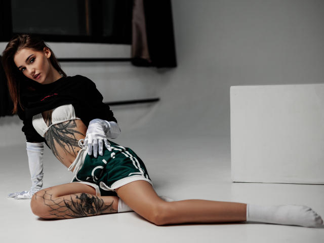 SophieKitty with tattoos, wearing a black crop top, green shorts, white gloves, and socks, laying on the floor, legs spread, showing her tits and ass.