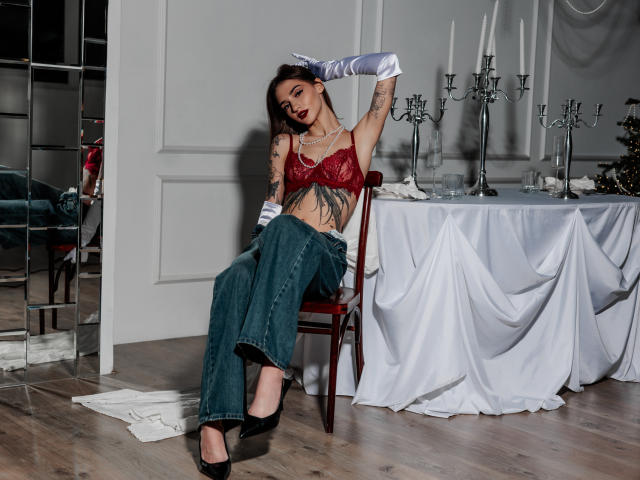 SophieKitty with tattoos, red lace bra, white gloves, green jeans, black heels, sitting on chair, arms raised, table with candles and white cloth in background.