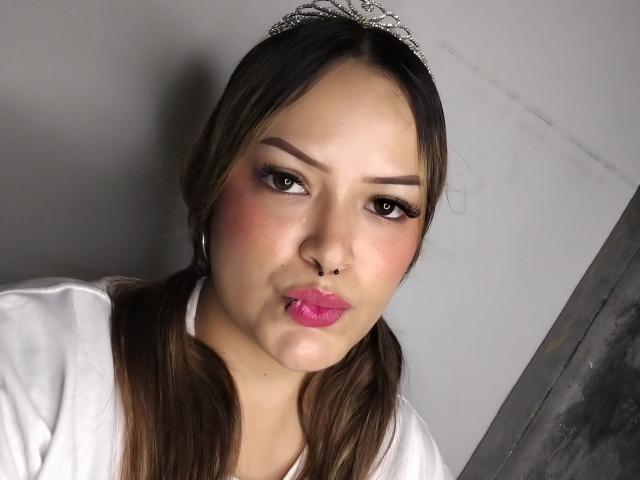 AllisonJustingb with pigtails, pink lipstick, and a septum piercing, pouting her glossy pink lips, wearing a white top and a jeweled tiara, brown eyes, and hoop earrings.