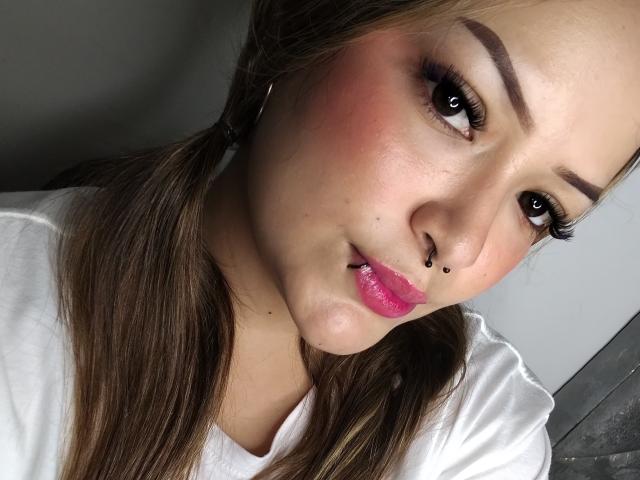 AlisonJustingb has puffy pink lips, a nose ring, brown pigtails, and pink blush on her chubby face, wearing a white shirt, with dark eyeliner and long lashes.
