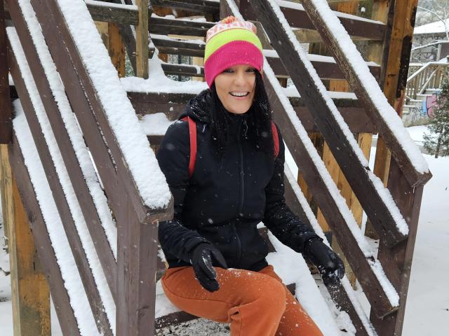 JoliDanielle with snow on wooden stairs, wearing a pink, yellow, and orange beanie, black jacket, orange pants, black gloves, smiling, snowflakes on her face and jacket.