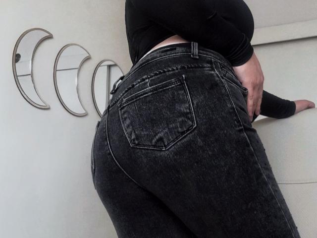 ValeriRose's ass in tight, dark blue denim jeans with visible pockets, wearing a black long-sleeve top, standing with one hand on a white wall.