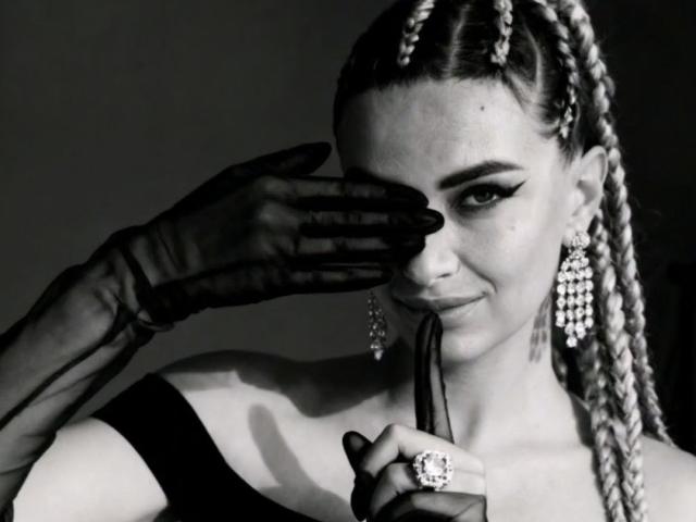 Black and white photo of PassionX with braided hair, wearing an off-shoulder top, black gloves, diamond earrings, and holding a ring, covering one eye with her gloved hand.