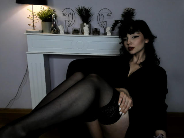 DeniseKitty with black hair, dark lipstick, and black lingerie, sitting with legs crossed, showing her black lace panties and sheer black stockings, arms resting on her thighs.