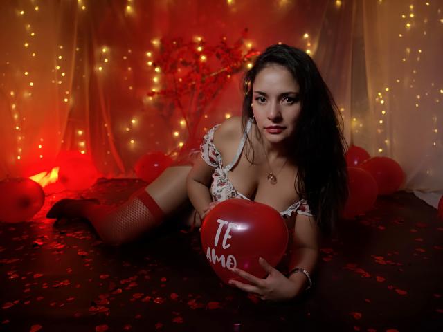 AngelaBranson with long dark hair, red fishnet stockings, floral bra, holding red balloon, laying on dark floor with red petals, red lights, looking at camera. Boobs, sexy.