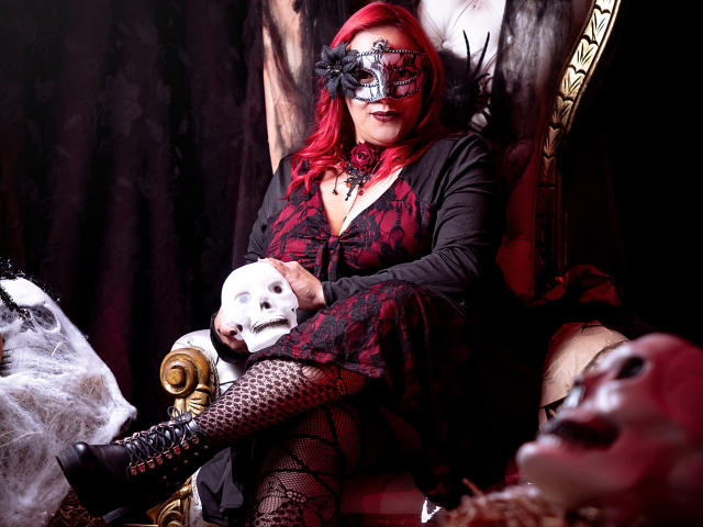 SammyOwens with red hair, black lace mask, red and black dress, fishnet stockings, black boots, holding white mask, sitting on ornate chair, surrounded by masks.