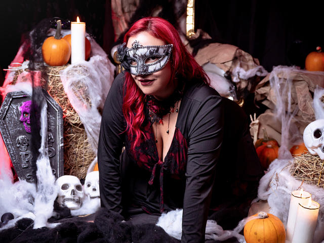 SammyOwens with red hair, black mask, and black outfit, kneeling on hay, surrounded by skulls, candles, pumpkins, and black fabric. Boobs visible under dress, looking sexy.