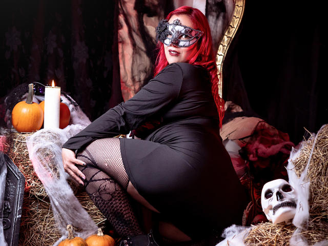 SammyOwens with red hair, black mask, black dress, fishnet stockings, and lace garters, kneeling on hay, showing her ass, surrounded by pumpkins, candles, and a skull.