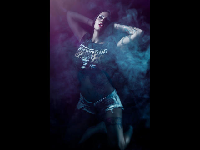 XANIA-WET, com tatuagens, vestindo uma cropped e shorts jeans, braços levantados, fumaça ao redor, iluminação em tons de roxo escuro e azul, batom vermelho, e uma pose confiante e sensual..
