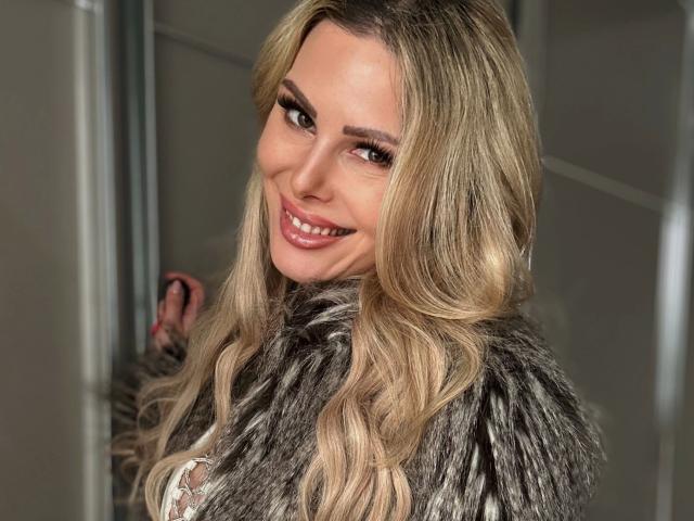 XANIA-WET, com longos cabelos loiros ondulados, sorrindo, vestindo um casaco cinza de pele, exibindo seus seios, tem um rosto bonito e sexy, e está olhando por cima do ombro..