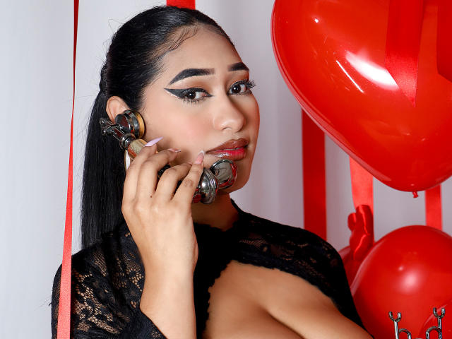 MelisaJackie with black lace top, holding metallic ball gag in mouth, red balloons, long black hair, thick eyebrows, red lipstick, large tits, pink nails, serious expression.