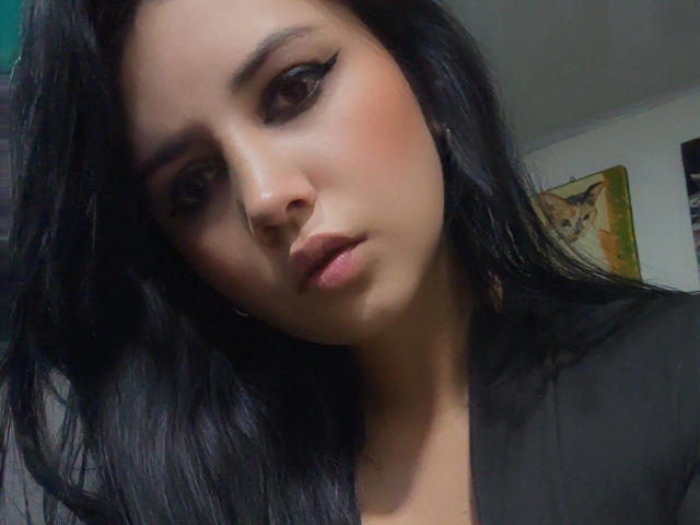 Kkatalina with dark, wavy hair, thick black eyeliner, and a serious expression, showing a small nose and full, pink lips, wearing a black top, looking slightly down.