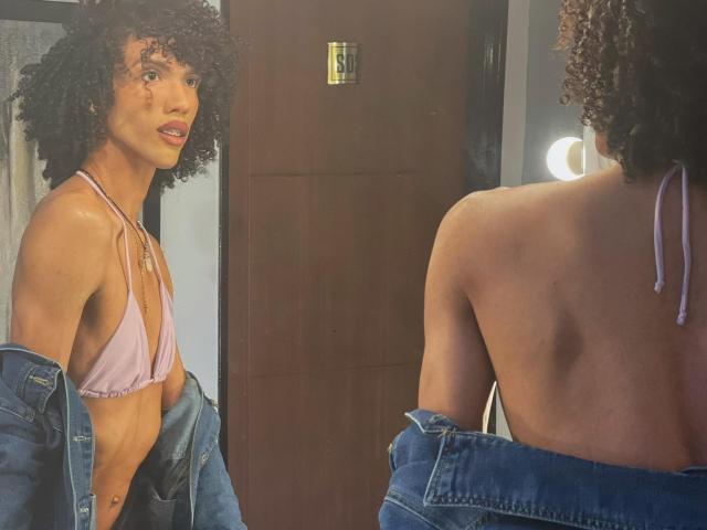 CamilleCedrik with curly hair, light brown skin, small pink bikini top, and blue jeans, looking in mirror, showing back with jeans down, revealing bare ass.