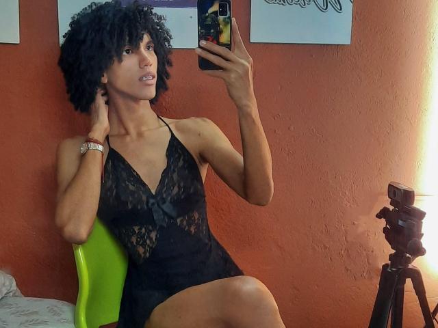 CamilleCedrik, avec ses cheveux noirs bouclés, portant une robe en dentelle noire, assise sur une chaise verte, prenant un selfie devant un miroir, montrant son corps mince et tonique et ses petites seins..