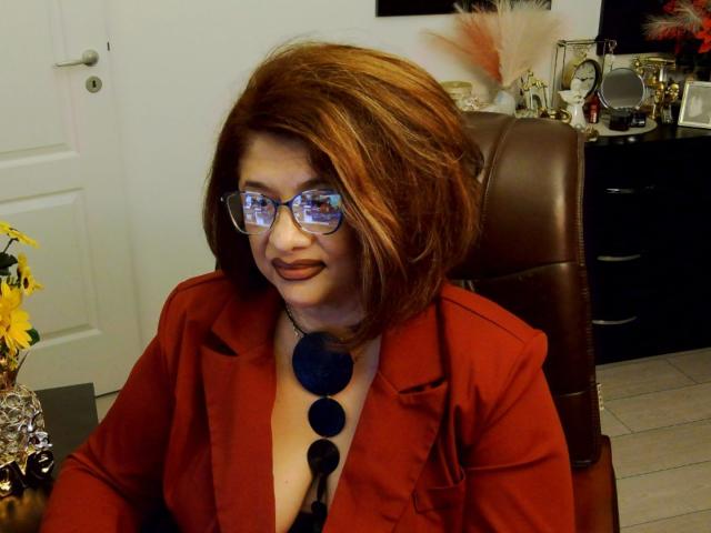 ClassybutNaughty with red hair, glasses, and a red blazer, showing cleavage with a blue necklace, sitting in a brown leather chair, smiling with brown lipstick.