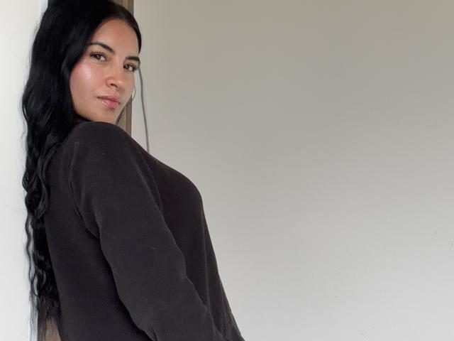 EmilyJensenn with long black hair, wearing a black sweater, showing her tits slightly, standing against a white wall, looking at the camera with a neutral expression.
