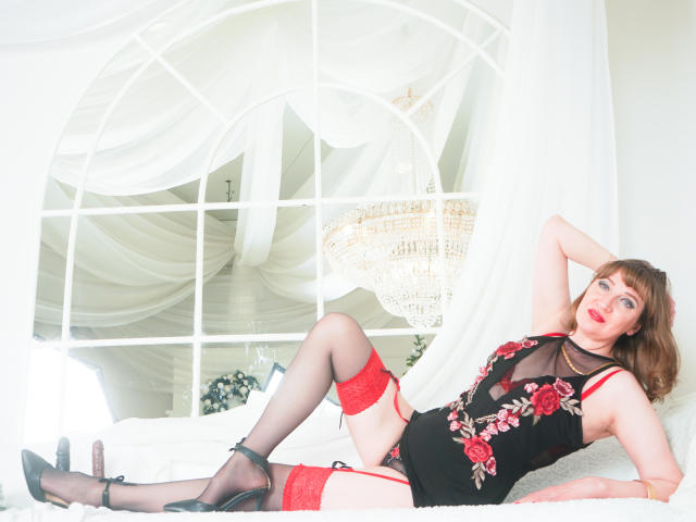 SquirtyCumShow lying on a bed, wearing black lingerie with red rose embroidery, red garter stockings, black heels, and red lace, her tits showing through the mesh, arms behind head.