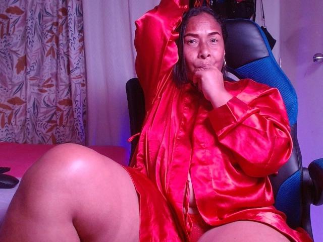HannaPasion, wearing a shiny red satin robe, sits with one arm raised, exposing her pussy, and her hand near her mouth, on a blue chair, looking confident.