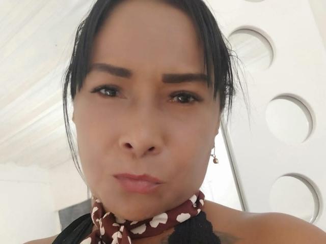 Close-up photo of HannaPasion with light brown skin, black hair pulled back, wearing a red halter top with white heart patterns. Background features a bright, metallic heart shape. She's pouting slightly.