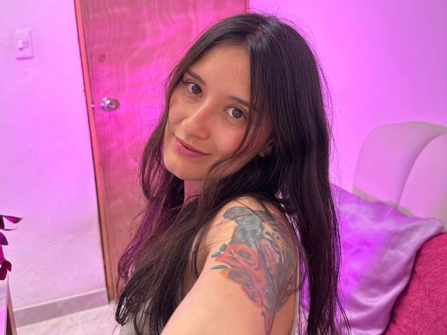 KimyRosse with long black hair, brown eyes, and a tattoo on her right shoulder, takes a selfie. She's looking at the camera with a subtle smile, in a pink-lit room. Fucking sexy.