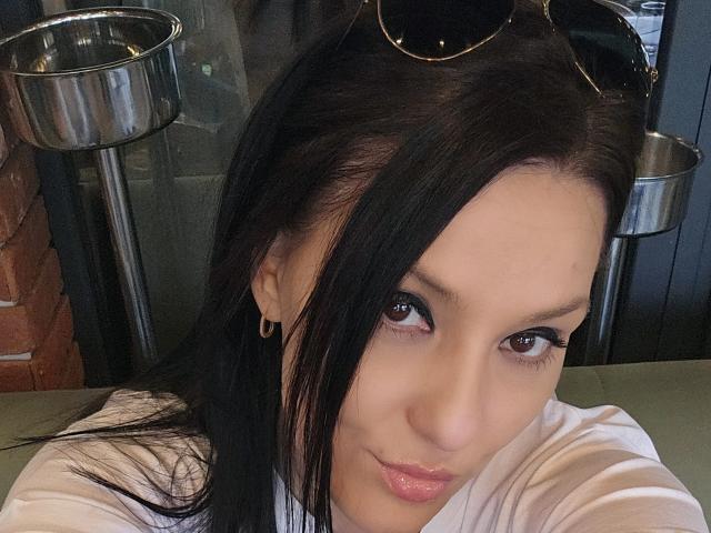 Close-up of JoliDanielle, a young woman with long black hair, wearing a white shirt, gazing seductively at the camera with glossy pink lips, metal kitchen pots in the background. Fucking sexy.