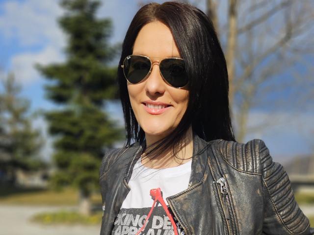JoliDanielle, a sexy woman with long black hair, wears dark sunglasses and a black leather jacket over a white graphic tee with a red ribbon. Background has blurred trees.