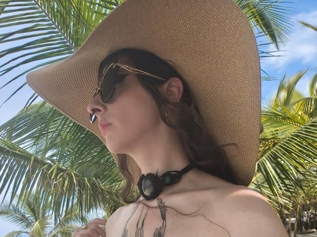 Photograph of KizzuKyuwu, a pale-skinned woman with dark brown hair, wearing a wide-brimmed straw hat and dark sunglasses, against a backdrop of vibrant green palm fronds. Her chest is bare, showing small breasts. She's wearing a black choker with a large pendant, and her eyes are half-closed. Sunlight highlights her features.