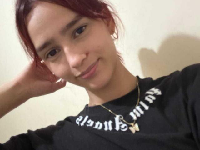Photograph of MelodyMansson with reddish-brown hair, smiling slightly, hand resting on head, wearing a black sweatshirt with white text, and a gold necklace with a cross pendant. Fucking cute.
